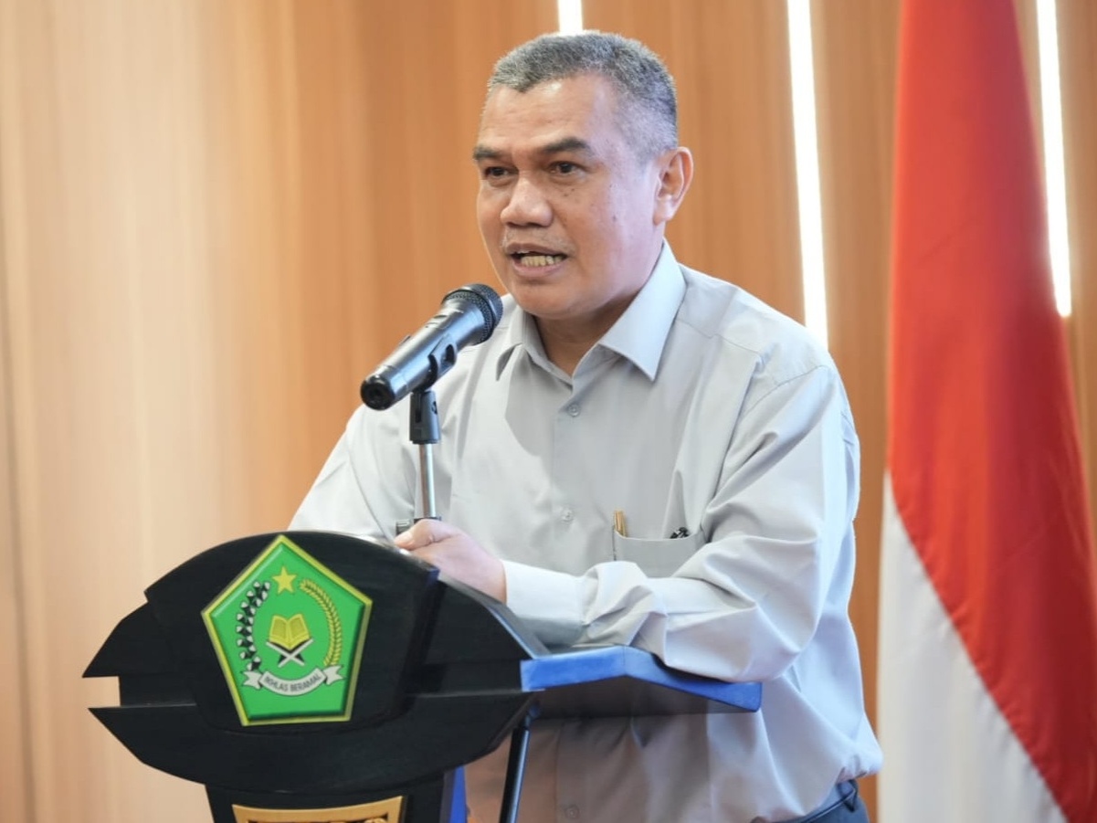 Inspektur Jenderal Kemenag Khairunas berbicara di podium dengan mikrofon, berlatar bendera Indonesia dan logo Kementerian Agama.