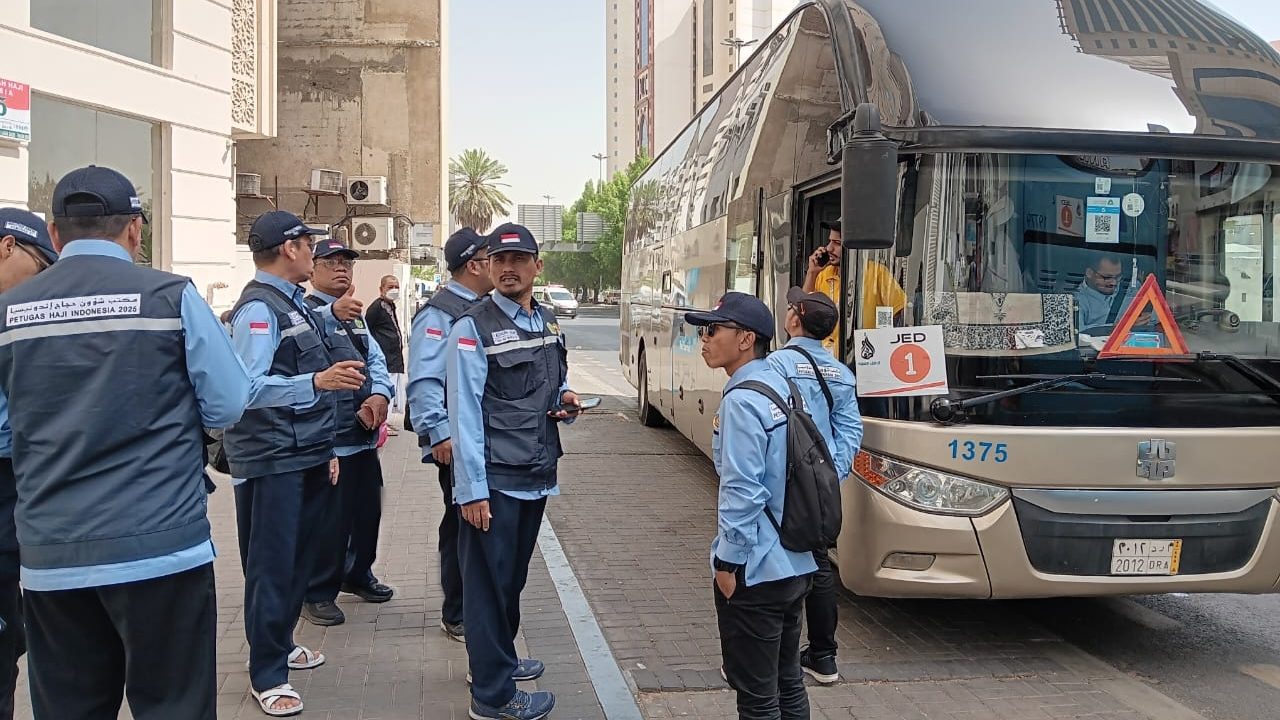 Petugas Haji Indonesia 2025 berdiri di dekat bus jemaah haji di Arab Saudi sambil berkoordinasi, mengenakan seragam biru tua dan rompi bertuliskan "Petugas Haji Indonesia 2025".