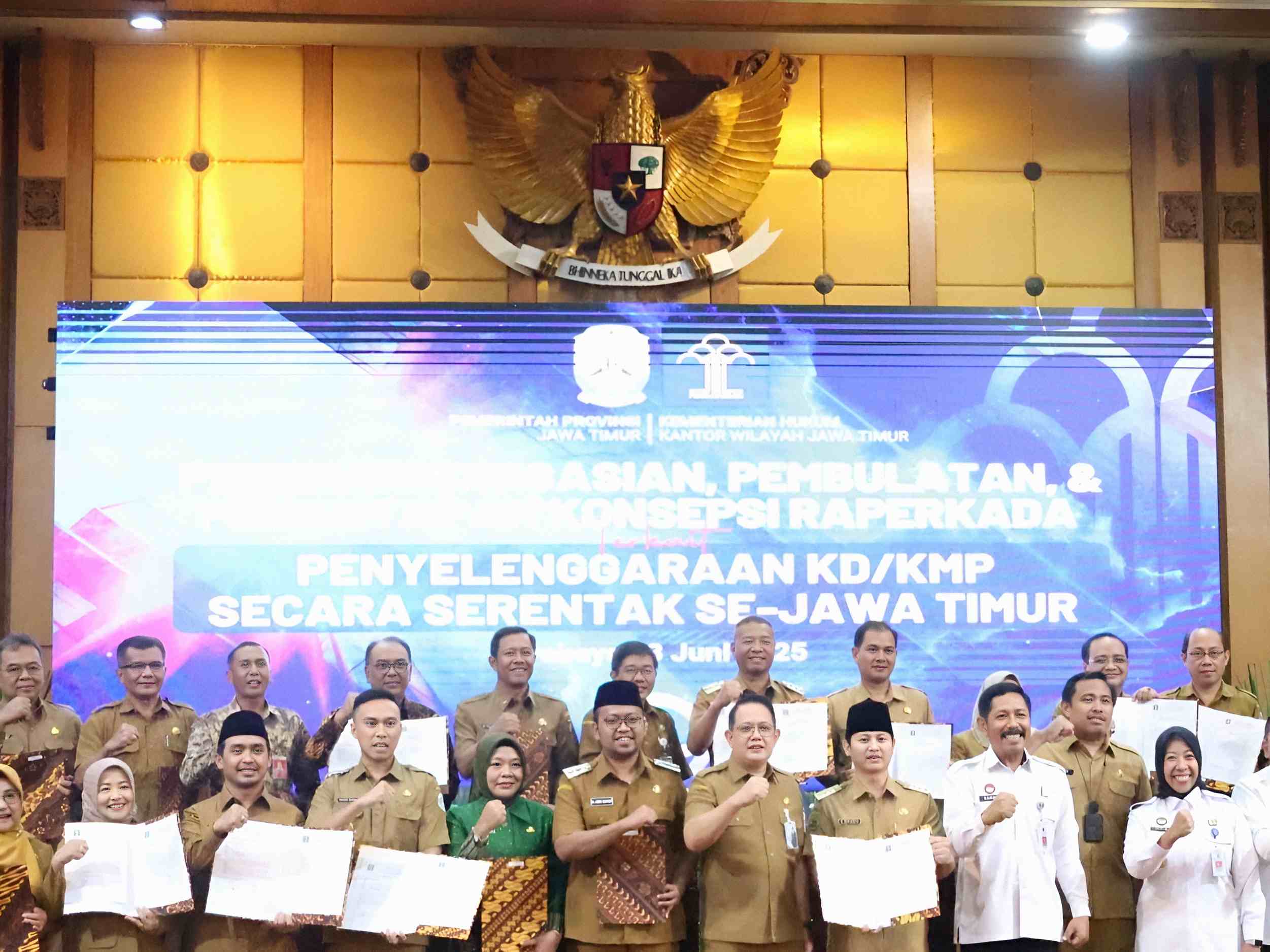 Wakil Bupati Sampang dan pejabat daerah berfoto bersama setelah penyerahan dokumen Raperkada KD/KMP se-Jawa Timur di Surabaya, di bawah latar belakang layar acara resmi.