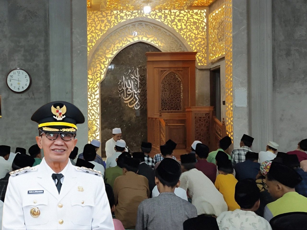 Bupati Pamekasan KH. Kholilurrahman berdiri mengenakan seragam dinas, di depan suasana pengajian kitab kuning di masjid pesantren bersama para santri.