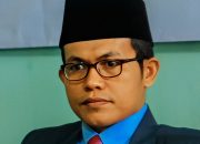 Pria berpeci hitam memakai jas dan dasi merah muda duduk di ruang formal dengan ekspresi tenang dan profesional.