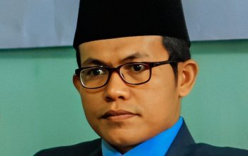 Pria berpeci hitam memakai jas dan dasi merah muda duduk di ruang formal dengan ekspresi tenang dan profesional.