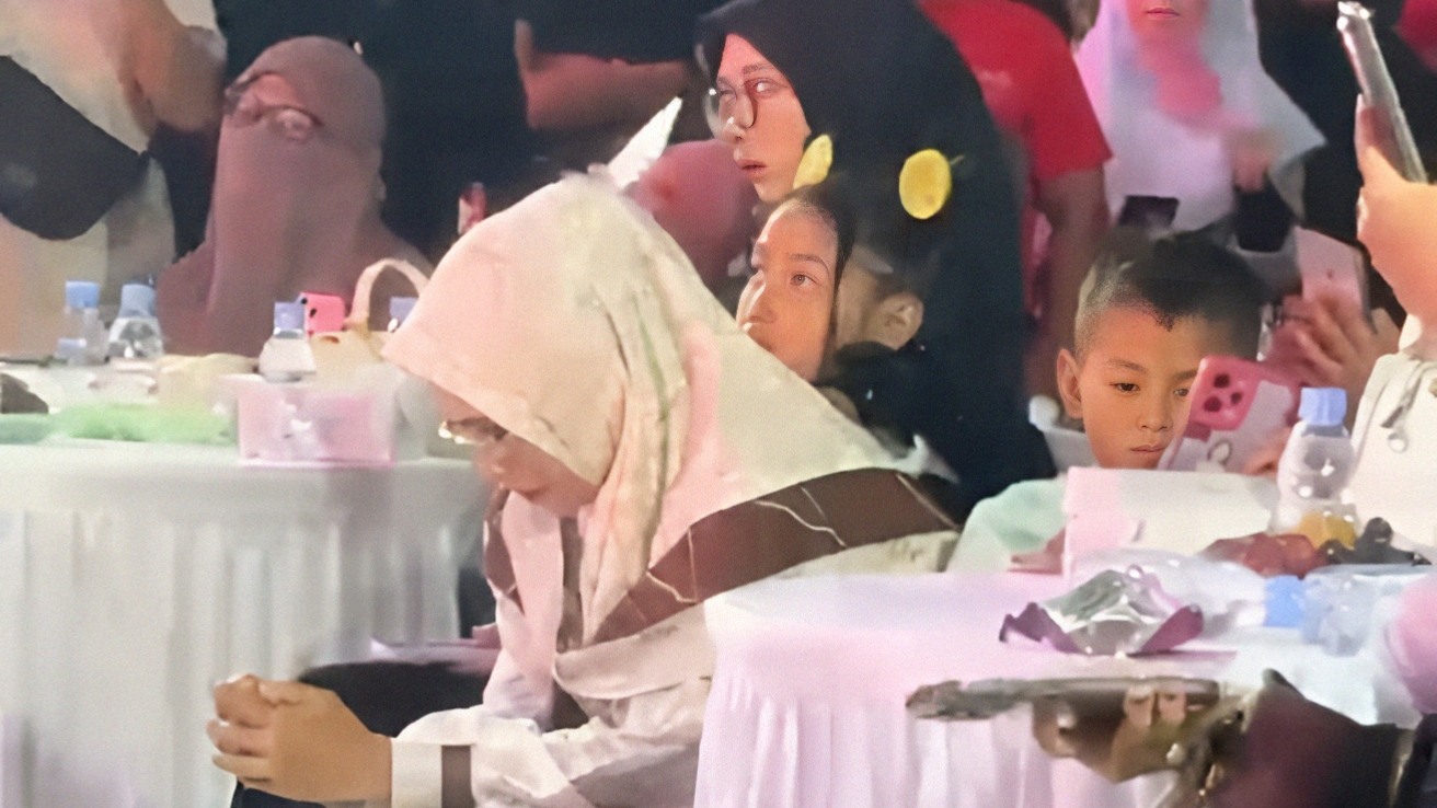 Seorang perempuan berhijab menunduk berdoa di antara penonton konser, dengan anak-anak dan orang lain duduk di meja sekitar.