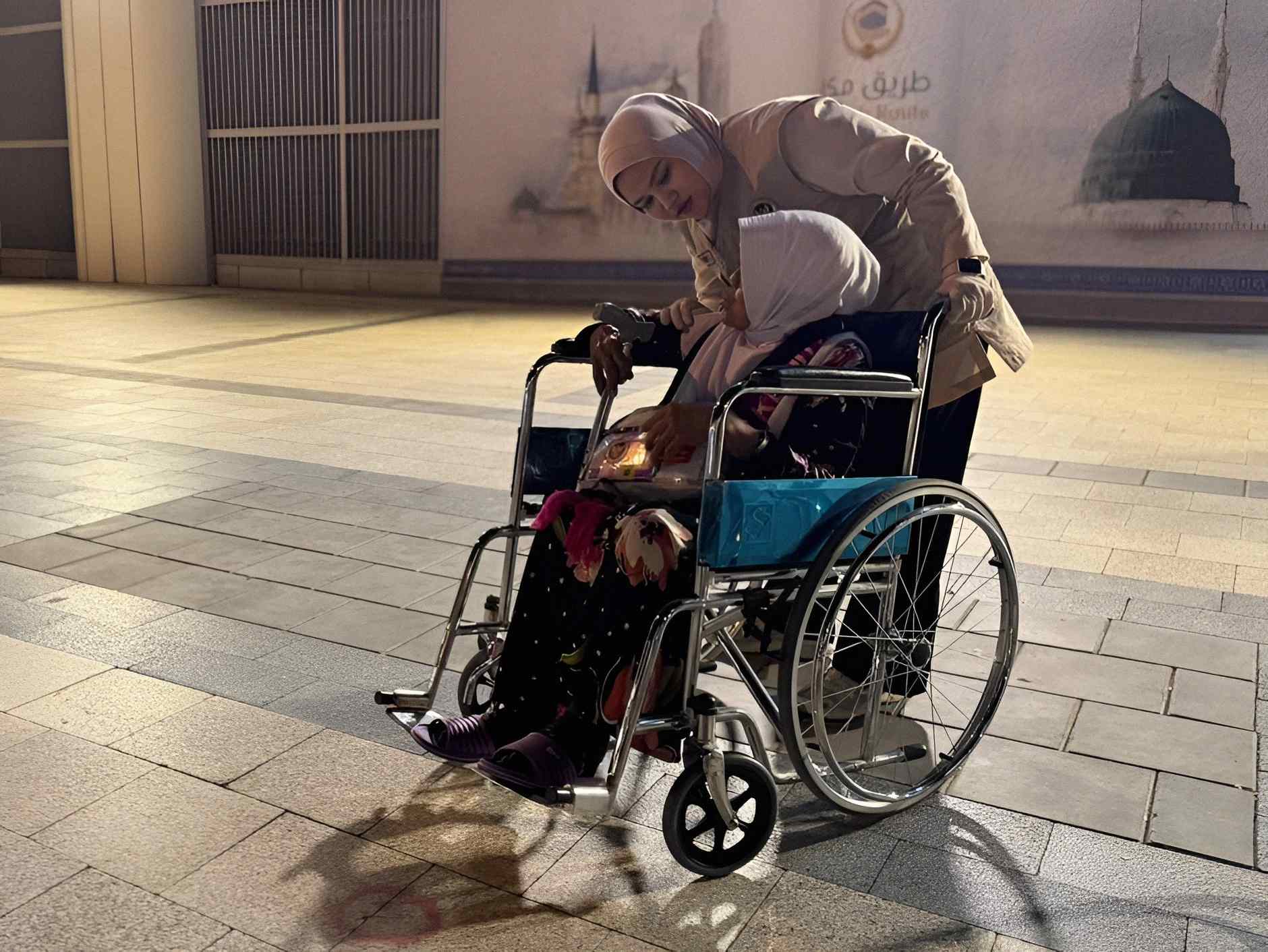Petugas haji mendorong jemaah lansia di kursi roda di area kedatangan Madinah pada malam hari.