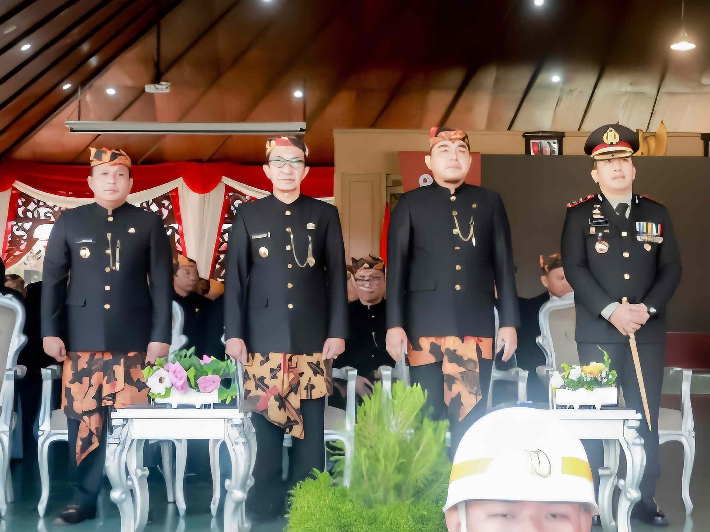 Empat pejabat mengenakan pakaian adat dan seragam dinas berdiri di area upacara, dengan peserta lain dan dekorasi merah putih di belakangnya.